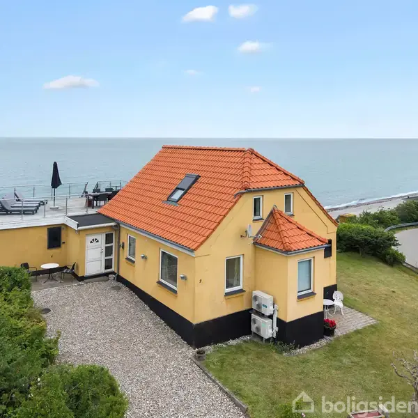 Gult hus med rødt tag, beboet ved havet med en grusbelagt indkørsel. Huset har en tagterrasse med parasol. Grøn have omkranser huset, og en vej snor sig langs kysten.