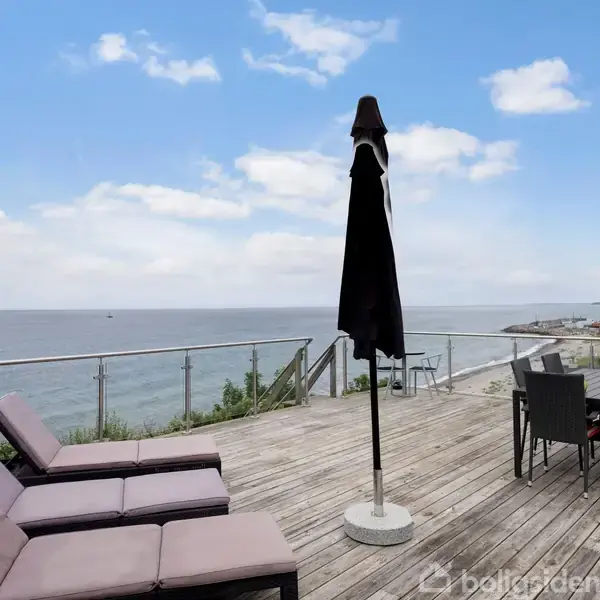 Terrasse med liggestole og parasol, roligt placeret mod en havudsigt. Til højre står et bord med stole, omgivet af glasgelænder, under en blå himmel med skyer.