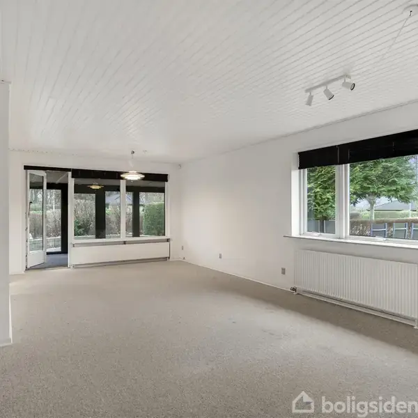 En tom stue med lyse vægge og tæppebelagt gulv. Store vinduer og en glasskydedør giver udsigt til haven. Loftet har hvid træpanel og en simpel lysekrone.