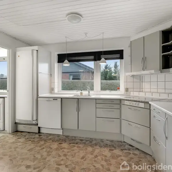 Køkken med grå skabe og skuffer, i hjørneopstilling, hvor en opvaskemaskine er integreret. Vinduet viser en udsigt til haven. Til venstre er der adgang til bryggerset.