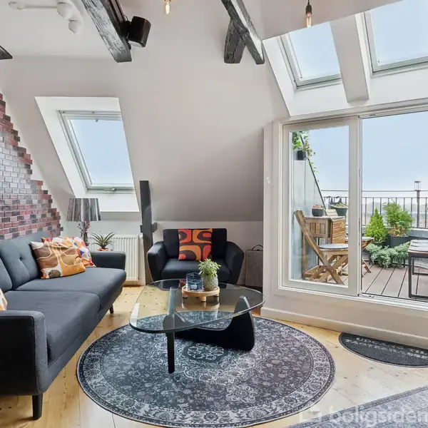 Sofa står i en stue med skråvægge og tagvinduer, omgivet af en kamin, tæppe og sofa. Gårdhave er synlig gennem glasskydedøre med træmøbler og planter på en terrasse.