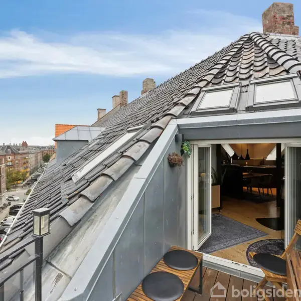 Tagterrasse med åbne døre fører til en hyggelig, moderne lejlighed. Omgivet af klassiske murstensbygninger i bykvarter. Små borde og stole står udenfor, under en klar himmel.