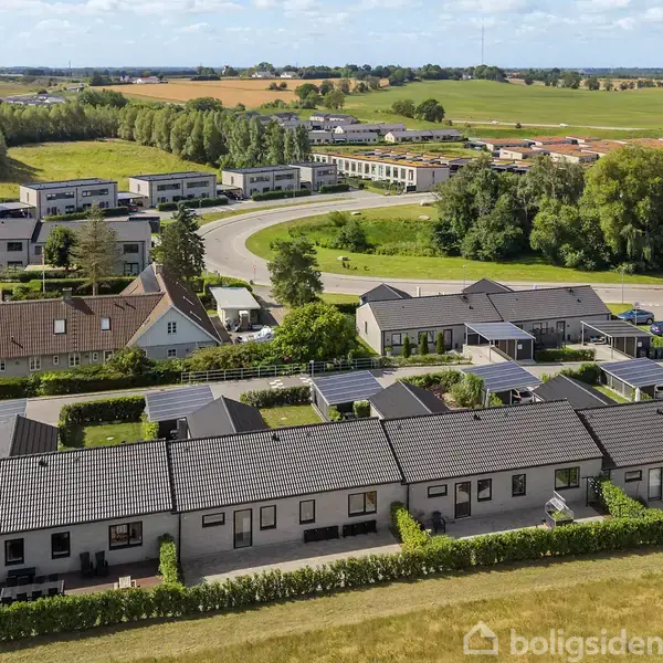 Rækkehuse med grå tage står på række i et landområde. Bag dem ses åbne marker, træer og moderne bygninger. Himmel med skyer giver en solrig atmosfære.