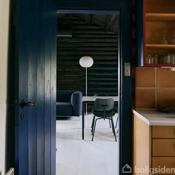 En åben dør åbner op til et rum med en sort væg, blå sofa, og sort stol ved bord. Baggrunden har en hvid gulvlampe. Køkkenbord og glasreol er til højre.