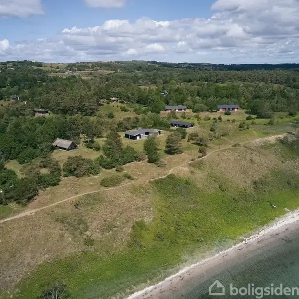 Kystlinje med græs og træer strækker sig langs havet. Flere huse ligger spredt på bakken, omgivet af skov og grønne områder under en delvist skyet himmel.