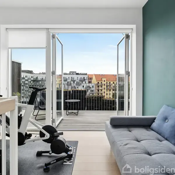 En lys stue med en grå sofa og træningsudstyr foran en åben altandør. Altanen har udsigt til en by med flere bygninger i baggrunden.