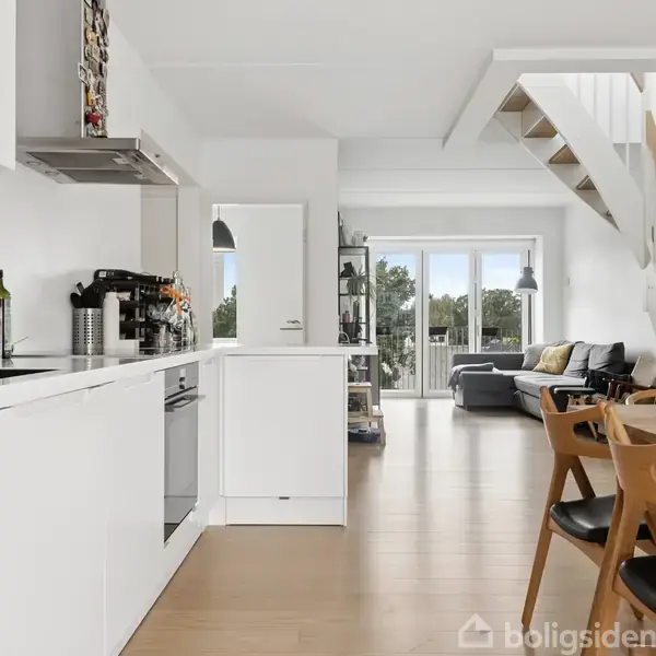 Et moderne køkken med hvide skabe åbner op til en stue, hvor der er en grå sofa nær vinduer. Spisebord med stole er i forgrunden. Trappe til højre.