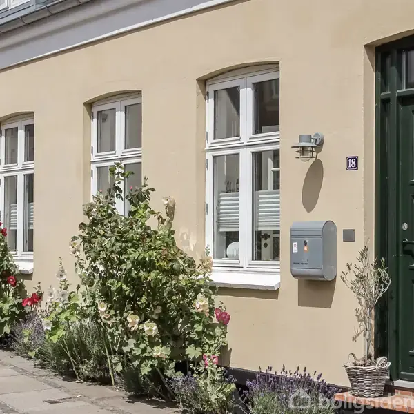 Et gult rækkehus med fire hvide vinduer og en mørkegrøn dør tildelt nummer 18, omgivet af blomstrende planter på fortovet i en rolig gade.