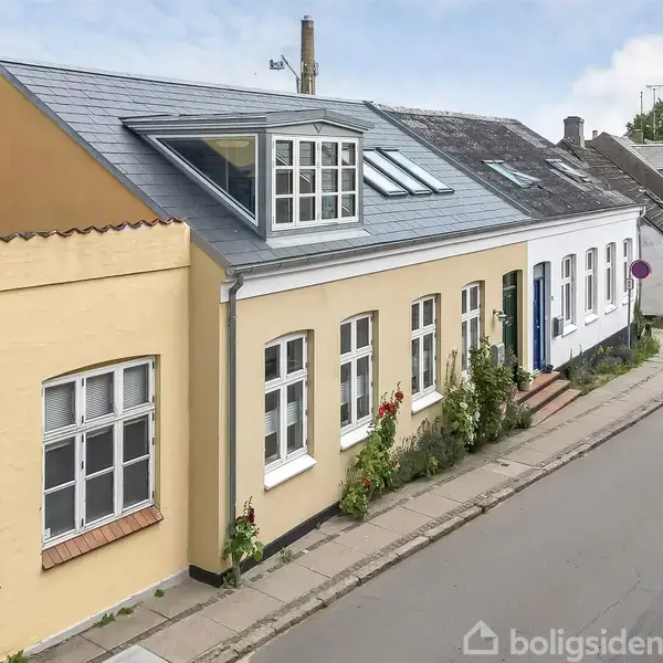 Et gult byhus med mørkt tag og tagvinduer, ligger langs en stille asfalteret vej. Foran huset vokser små buske og blomster, mens en rød bil er parkeret til højre. Nummeret "20" er synligt på væggen.