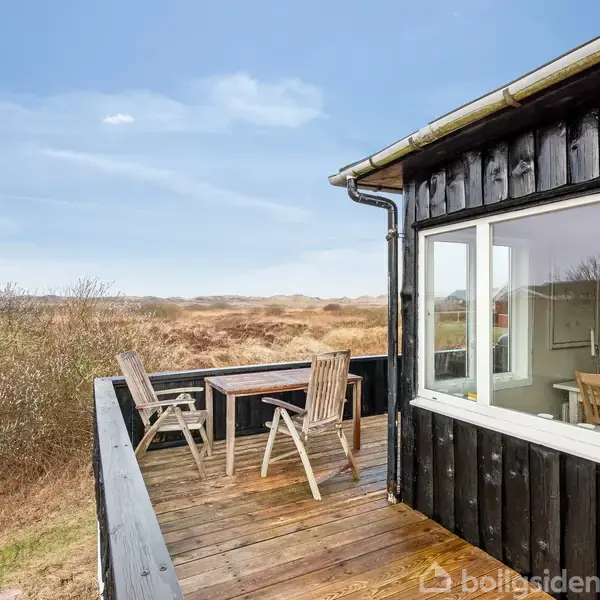 En terrasse med to træstole og et bord står udendørs. Huset har sorte trævægge og store vinduer. Omgivet af en landskab udsigt med buske og åbent land.