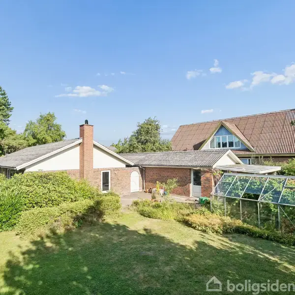 En etplansvilla med en glasudestue står i en frodig have med græsplæne og buske. De omkringliggende træer danner en grøn ramme under en klar blå himmel.