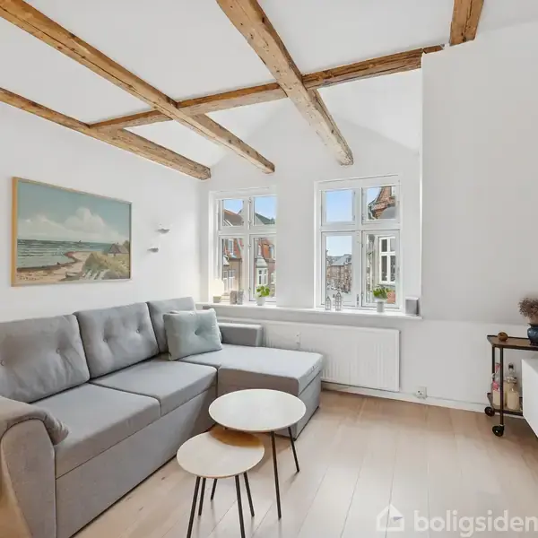 Grå sofa i en lys stue, placeret under et skrå loft med synlige træbjælker. To runde bord i front. Vinduerne viser en byudsigt. Vægmaleri til venstre.