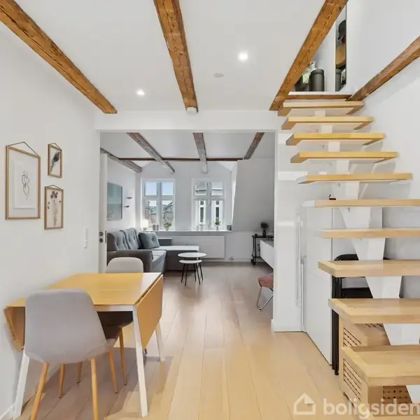 Et moderne rum med en trappe af træplanker, der fører til en overetage. Til venstre står et spisebord med to stole. I baggrunden er der en stue med sofa og vinduer. Træbjælker pryder loftet.