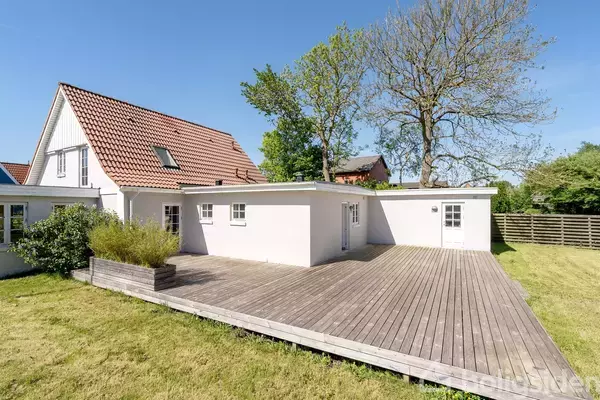 Et hvidt hus med rødt tag står i haven. Foran huset er en stor træterrasse. Omgivet af grønne planter og et hegn i baggrunden.