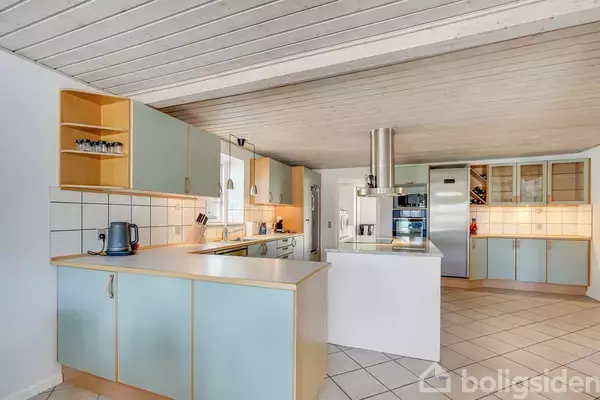 Et moderne køkken med lyseblå skabe og ståludstyr. En elkedel står på bordet, og der er hvide fliser på væggen. Omgivet af beige flisegulv og træloft.