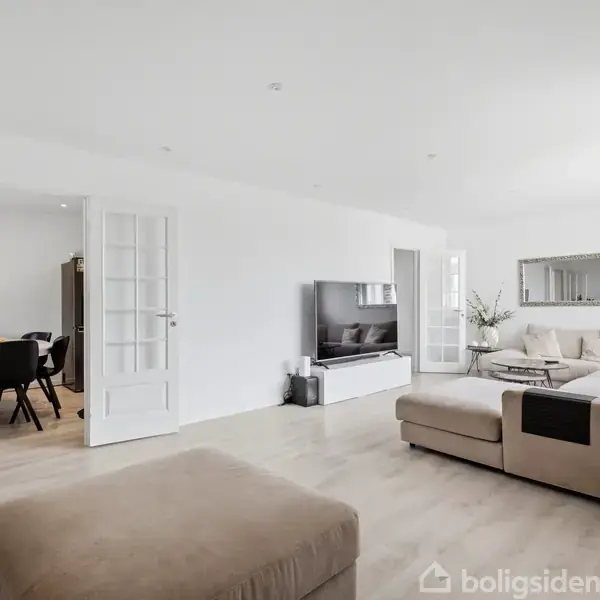 En lys stue har en beige sofa og TV, mens en spisestue med sorte stole ses gennem franske døre. Væggene er hvide, og gulvet er lyst trægulv.