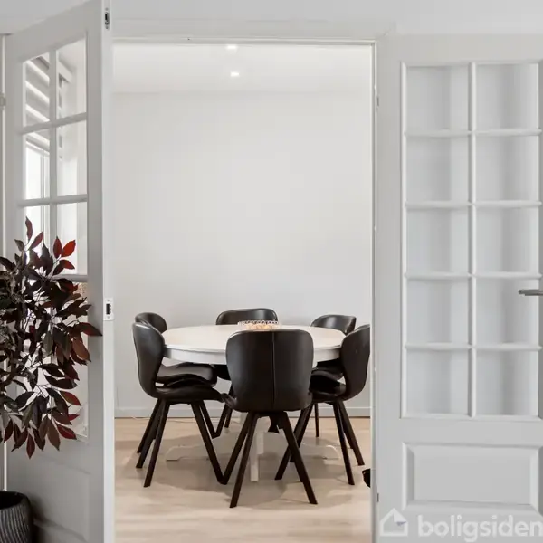 En rund spisebord med seks sorte stole står i et lyst rum. Glassdøre åbner ind til rummet, hvor en plante er placeret ved siden af døren.