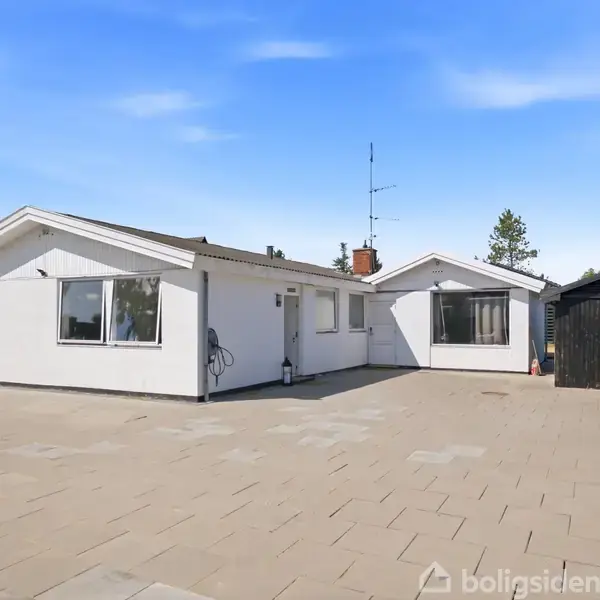 Hvidt, fladt hus står stille foran en stor brolagt gårdsplads. Ved siden af huset er en sort træbygning. Himlen er klar blå med få skyer.