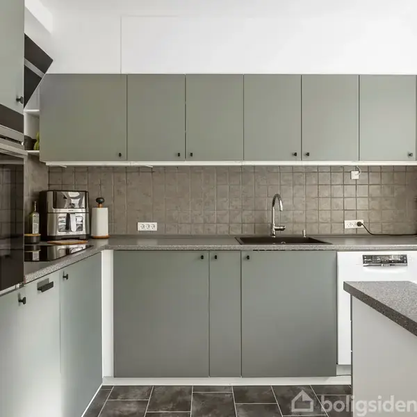 Køkkenskabe i lysegrønne nuancer optager rummet. Overfladerne har grå fliser, og rummet rummer en vask, kogeplader og forskellige køkkenapparater. En kaffemaskine og en elkoger står til venstre.
