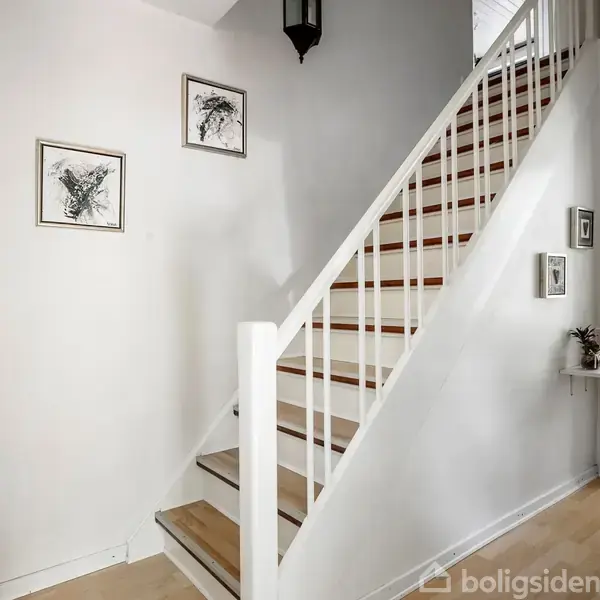 En trappe fører opad, omgivet af lyse vægge med hængende billeder. Til højre er en åben dør ind til et rum med flisegulv og moderne indretning.