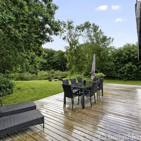 Et sommerhus står på en grøn græsplæne, omgivet af høje træer. Terrassen har havemøbler og store potteplanter. Ved siden af ligger et lille, sort udhus.