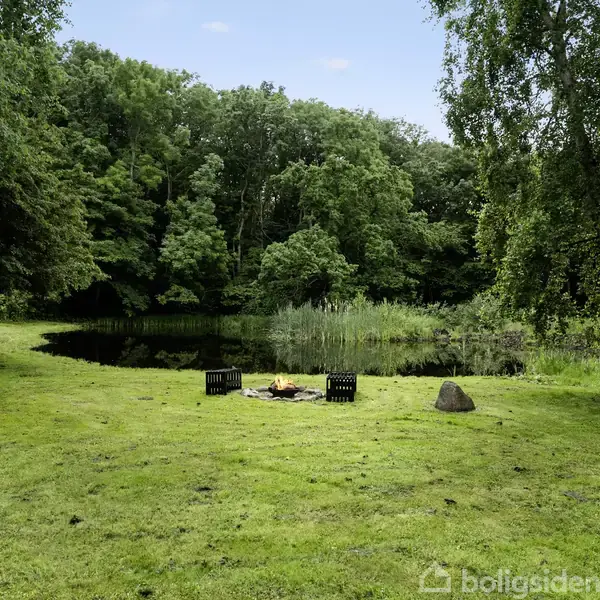 Et træhus står stille omgivet af tæt skov. En stor træterrasse med havemøbler strækker sig fra huset. Græsplænen er velplejet, og et lille skur ses til venstre.