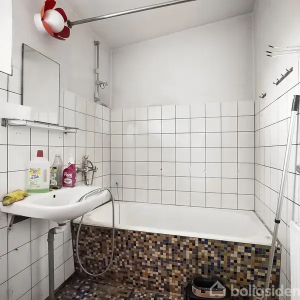 Badeværelse med badekar, omgivet af kvadratiske hvide fliser. Vasken har hylde med rengøringsartikler over. Vindue øverst, med rød hvid lampe. Gulvfliser i mønster, bruseforhængsstang over karret.