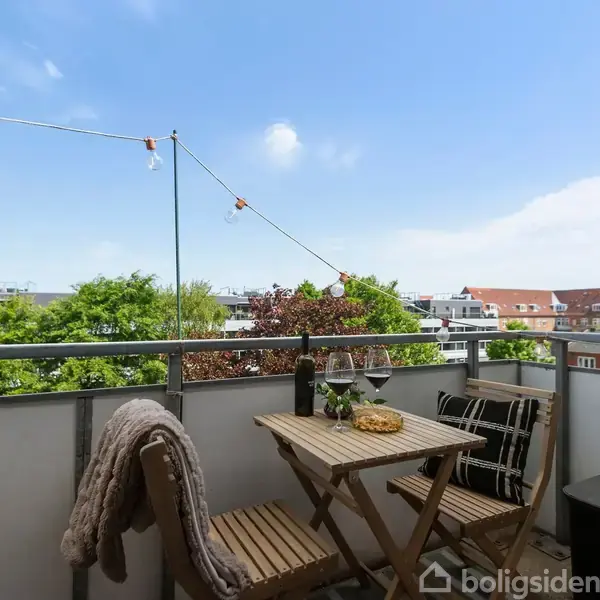 Et lille altanområde med et bord og to stole, pyntet med blomster og vin. Lyskæde hænger overalt, omgivet af grønne træer og byhuse i baggrunden under en blå himmel.