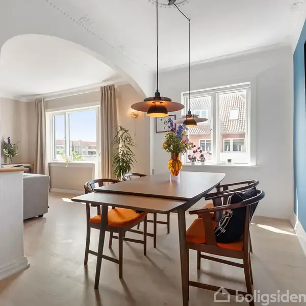 Spisebord med fire stole står i et lyst rum. Der er orange hynder på stolene og en lampe hænger over bordet. Rummet har vinduer med udsigt til nabotage.