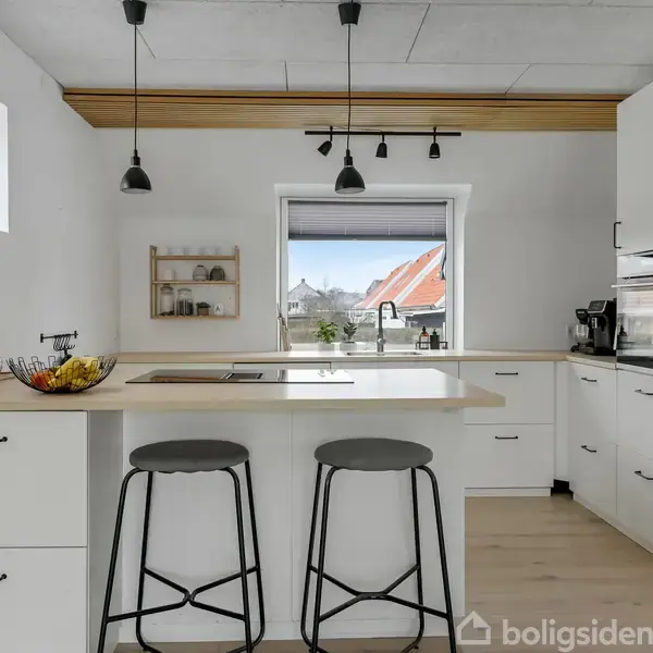Moderne køkken med hvidt interiør, der har en kogeø med to barstole. Tre hængelamper lyser op. Vinduet viser udsigt til røde hustage. Skuffer og skabe omgiver rummet.