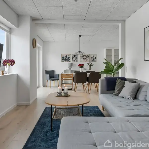 En stue med en grå sofa og et rundt sofabord står på et blåt tæppe. Et fladskærms-tv hænger på væggen. I baggrunden ses et spisebord med stole. Vaser med blomster er placeret nær vinduerne. Væggene har minimalistisk kunst.