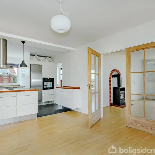 Et rummeligt, lyst køkken med hvide elementer og en kogeø. Dobbelte glasdøre åbner mod en træbeklædt stue med pejs. Store vinduer. Enkelt, moderne indretning med neutral farvepalet.