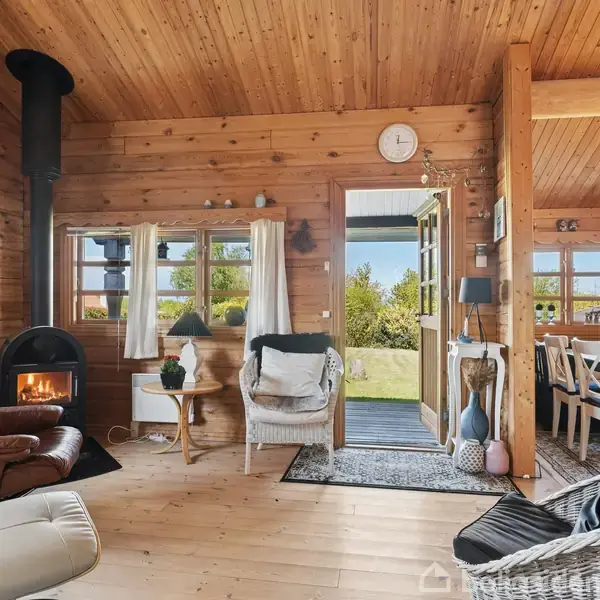 Pejselounge med brændende ovn, lænestole og tæpper, trævægge og loft. Døre åbner til grøn have, spisestue med dækket bord til højre, lysindfald fra store vinduer.