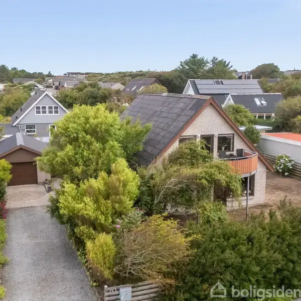 Et gråmalet hus med sort tag står omgivet af frodig have, tæt på huse i et fredeligt kvarter. En grusvej fører op til huset, der har en træterrasse.