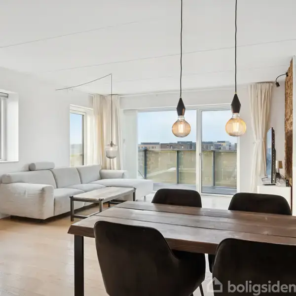 En lys stue med en grå hjørnesofa og sofabord til venstre. Træbord med fire sorte stole i forgrunden. Store vinduer og glasdør fører ud til en balkon.