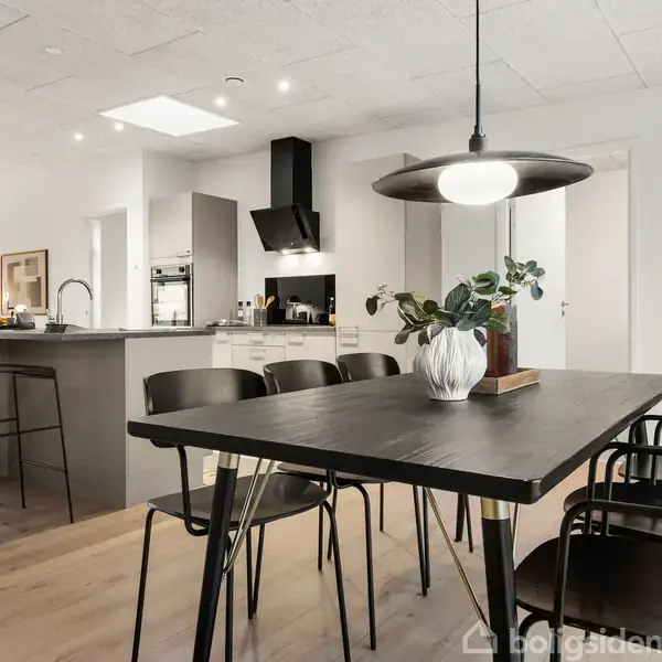 Et moderne køkken-alrum med et spisebord i forgrunden dekoreret med en plante i en vase. I baggrunden ses et køkken-ø med barstole, ovn og emhætte.