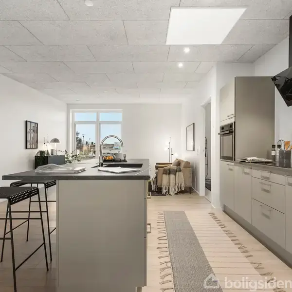 Et moderne køkken med en ø står i midten, omgivet af barstole. Køkkenet har indbyggede apparater og en emhætte. Lyst, med udsigt til vinduer og adgang til stue.