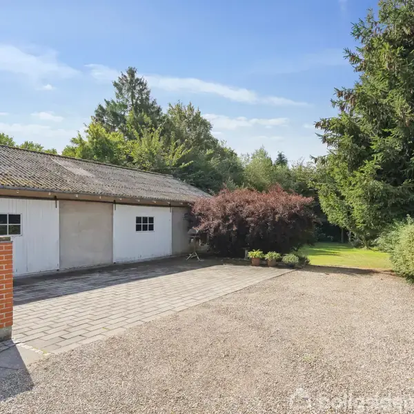 En garagebygning med hvide døre står ved en grusbelagt indkørsel. Omkring er der grønne buske og træer under en klar blå himmel.