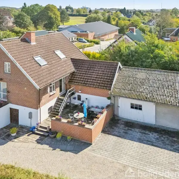 En murstensbygning med tagvinduer ligger i et landområde. En trappe fører til en hævet terrasse med havemøbler. Omgivet af grøn beplantning og flere huse i baggrunden.