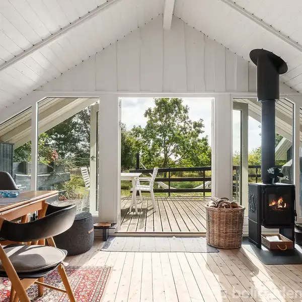 Træbeklædt stue med spisebord og stole står foran glasdøre, der åbner ud til en terrasse med udsigt til træer. En brændeovn står til højre ved siden af en kurv.