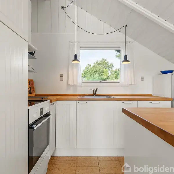 Et hvidt køkken med træbordplader omfatter en håndvask og en indbygget ovn. To hængelamper lyser området op. Et vindue viser udsigt til træer udenfor.