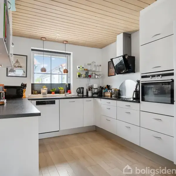 Et moderne køkken med hvide skabe og sorte bordplader. Apparater som ovn og mikrobølgeovn er indbygget. Vinduet centralt over vasken giver naturligt lys. Trægulv og træloft skaber en varm atmosfære.