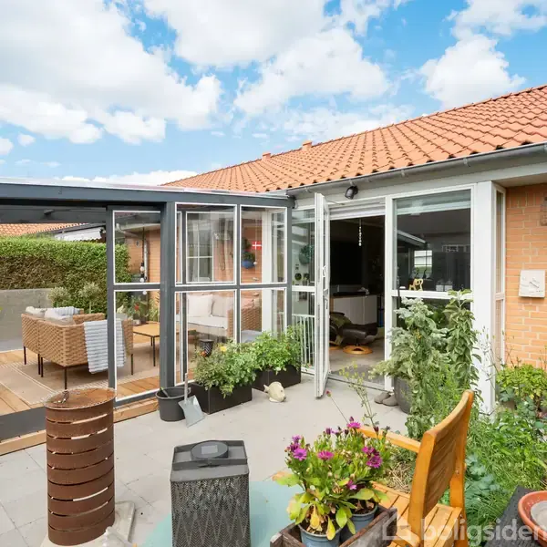 Et orangeri med glasvægge er fyldt med planter og åbner ud til en hyggelig terrasse. I baggrunden ses en rød murstensbygning med et rødt tegltag under en skyfri himmel.