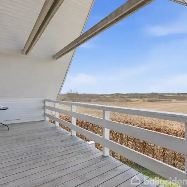 En balkon med trægulv og hvidt rækværk har et lille rundt bord mod en murstensvæg. Balkonen vender ud mod en åben, bakket landskabshorisont under en klar blå himmel.