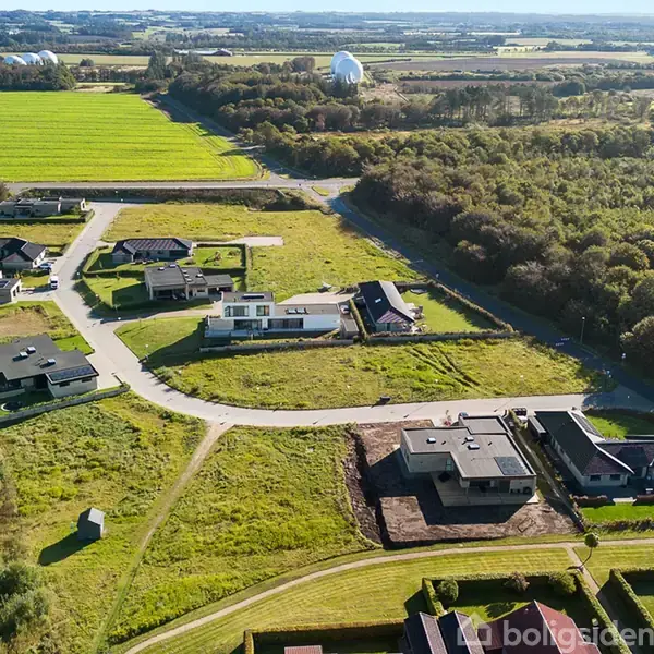 Boligområdet med moderne huse ligger i et grønt landskab. Veje forbinder bebyggelsen, mens marker og skove omkranser bygningerne, skabende en rolig, landlig atmosfære.
