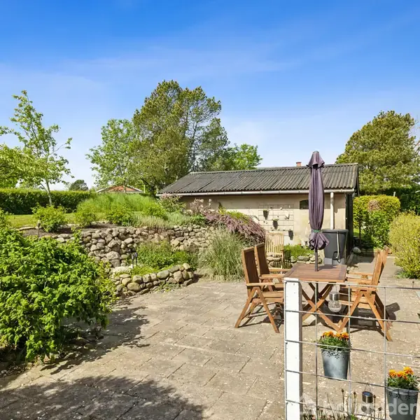 Træmøbler står på en terrasse med parasol, omgivet af en stenmur og frodig have. En bygning i mursten ses til højre, og himlen er klar blå.