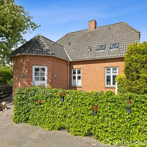 Et rødt murstenshus står i haven omgivet af grønne buske og træer, under en klar blå himmel. Huset har flere vinduer, og tagstenene er grå.