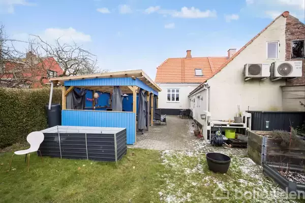 Et udendørs område med en lille blå beboelsesbygning til venstre. Et hvidt hus med rødt tag ses bagved. Haven har græs, en hvid stol og en hævet plantekasse.
