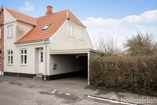 Et to-etagers hus med rødt tegltag ligger ved siden af en garage, omgivet af en hæk. Huset har hvide vægge og et lille indgangsparti ud til en gade.