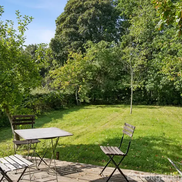 Et bord og to stole står på en terrasse. Sollys oplyser terrassen, mens grønne træer og en græsplæne omringer området og skaber en fredfyldt have.